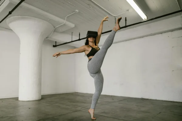 A woman performs a high kick while wearing a VR headset in an industrial indoor space.