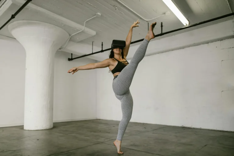 A woman performs a high kick while wearing a VR headset in an industrial indoor space.