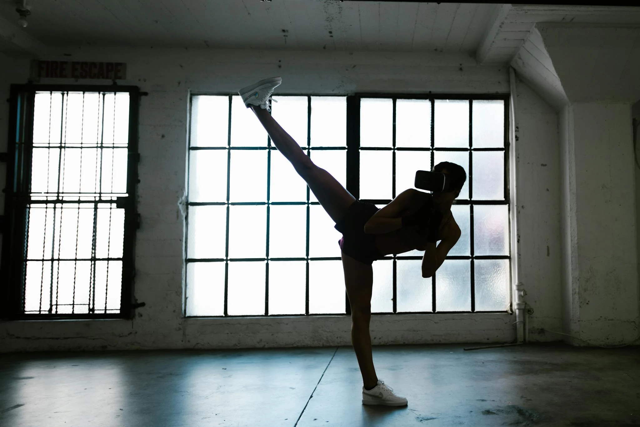 Silhouette of a woman wearing VR headset practicing high kick in a modern studio setting.