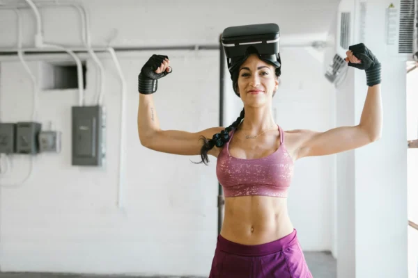 Woman flexes muscles with VR headset and gloves in gym, showcasing fitness technology.